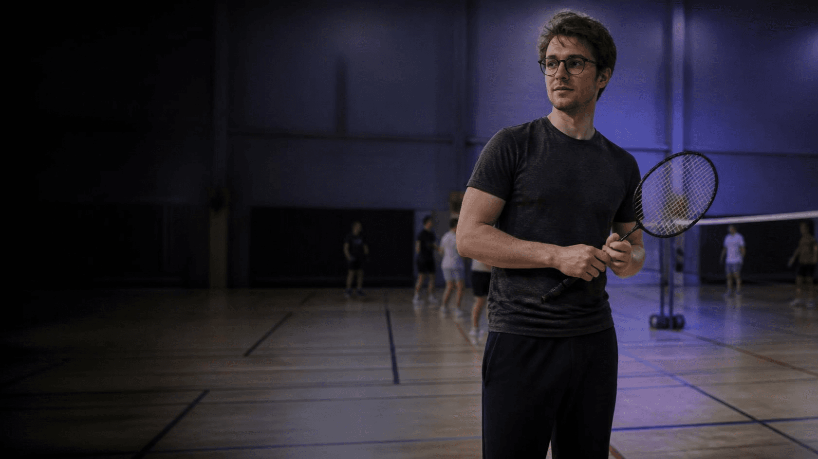 Un jeune homme tient une raquette de badminton au bord d'un terrain, lors d'une séance découverte dans un gymnase.