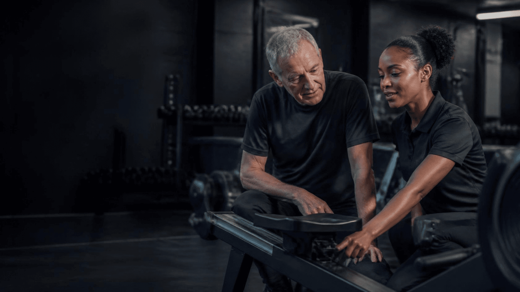 Dans une salle de sport calme, une employée montre à un homme aux cheveux gris comment régler un rameur.